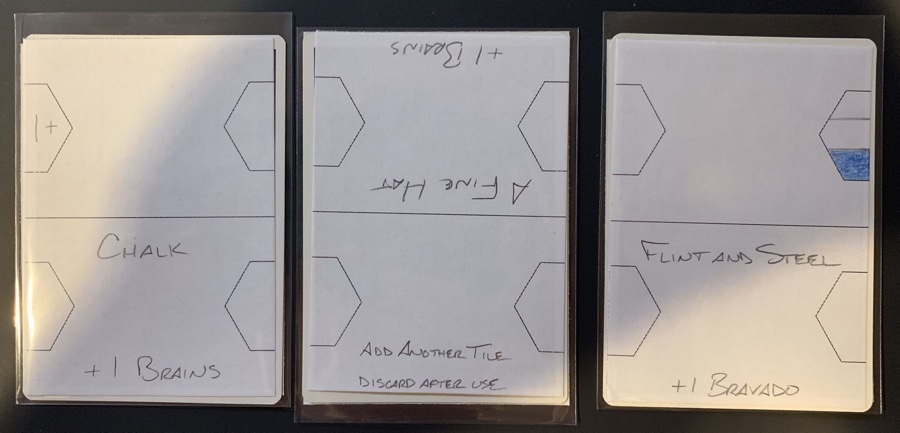 Three starter cards: Chalk, giving +1 Brains, has a +1 right gem on the other side; A Fine Hat, also giving +1 Brains, has a currently unnamed item on the other side which allows you to add another tile; and Flint and Steel, giving +1 Bravado, has a Brains left gem on the other side.